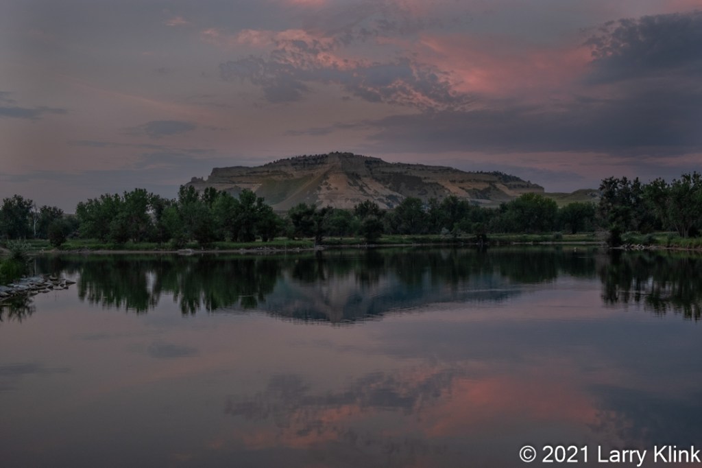 Image of Scottsbluff, a flat top butte, with the North Platte River in the foreground taken at sunrise. The clouds and their reflection in the river carry the magenta color of early sunrise.