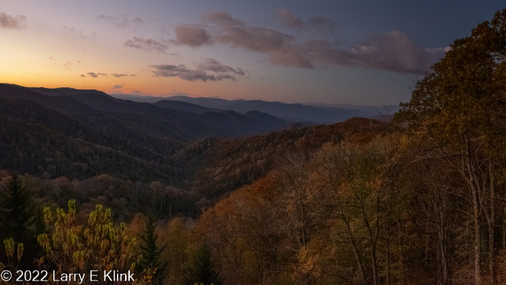 A mountain valley at early sunrise. The sun is casting an orange-yellow glow against a washed-out blue sky dotted with gray clouds. The mountain valley is forested. The trees are dark but adorned with autumn red, rust, orange and yellow.
