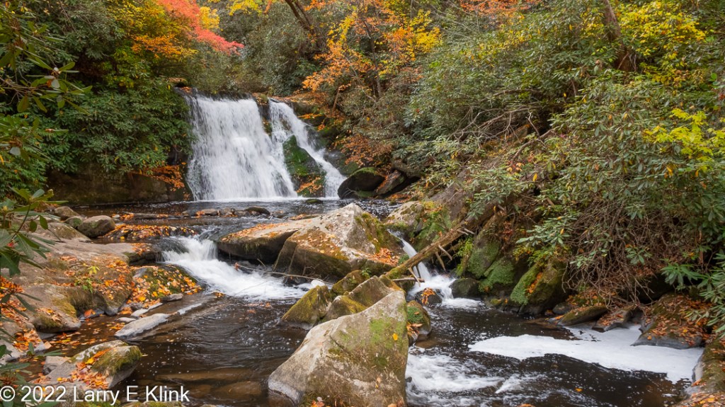 Image of a waterfall in Autumn. The waterfall is white, broken by some green. The white river projects from the bottom of the water fall, widening to the bottom of the frame. The boulders along and inside the river are light gray and strewn with golden brown and yellow leaves as well as some green moss. There is green foliage along the side of the river and some green, red and orange foliage above and to the back of the waterfall.