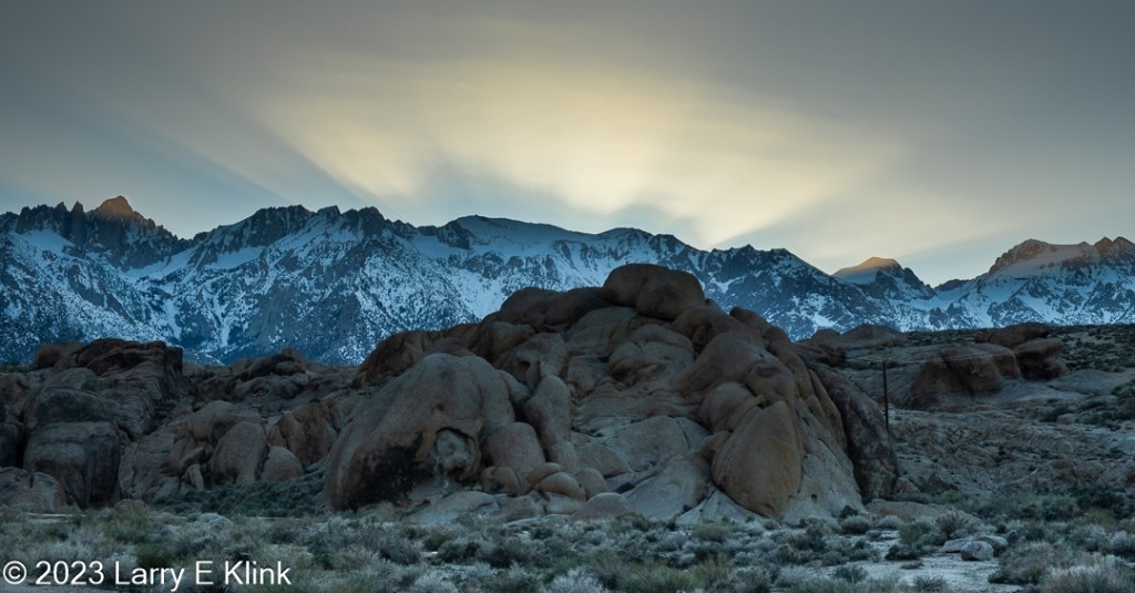 A photograph of the Alabama Hills in Eastern California at sunset.  The foreground of the photo is a desert landscape with clumps of green foliage on gray-brown and white sand. To the left side is a large mass of brown boulders of irregular shape. There are more boulders, foliage, and gray-brown soil to the right of the boulders. The middle ground is the snow-capped peaks of the Sierra Nevada mountains. In this picture they appear blue with white snow. One of the peaks on the left and two on the right are gold, highlighted by the sun. The sky is light blue. A large portion of the sky is highlighted by thin clouds that are creamy gold colored, caused by the reflection of the sun.