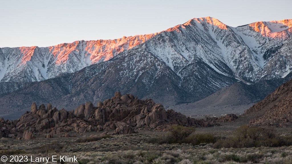 A photograph of a portion of the Sierra Nevada mountains, at sunrise, from the desert valley of the Alabama Hills of Eastern California.  The foreground is the desert landscape of the valley. It is mostly clumps of green foliage on tan soil. The left two thirds of the foreground are brown clumps of irregular boulders tapering to the ground on their right side. To their right of the boulders is a slope of brown boulders with soil at its base. The middle ground is the snow-capped mountains. Their base is dark brown, mottled with snow and some nearly indistinguishable dark clumps of foliage. The peaks are a red orange, mottled with snow. This color is the Alpen glow as the rising sun paints the top of the peaks. The upper section of the image is the blue sky.
