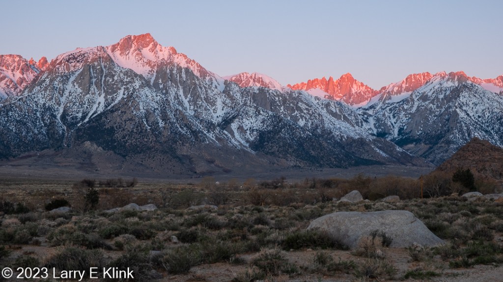 A photograph of a portion of the Sierra Nevada mountains at sunrise from the desert valley of the Alabama Hills of Eastern California.  The foreground is the desert landscape of the valley. It is mostly clumps of green foliage on tan soil. It is strewn with gray boulders. On the right, set further back, is a steep slope of brown boulders. The middle ground is the snow-capped mountains. At their base is the dark brown alluvium eroded from the mountains which is dotted with dark spots of foliage. Rising, the mountains are mostly blue, mottled by white snow. The peaks are a red orange, mottled with snow. This color is the Alpen glow as the rising sun paints the top of the peaks. The upper section of the image is the blue sky.