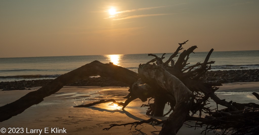 Picture of the sun, mid-level on the horizon, silhouetting dead trees and brush of Driftwood Beach, while illuminating the ocean and the sky. The sky in the background is golden gray at the top that turns more gold as it flows to the bottom. The sun is a watery yellow circle left of the middle, near the top, of the picture. The foreground is the light tan sand of the beach. The ocean is gray with a strong golden reflection down the middle. Rocks separate the beach from the ocean. The beach in the foreground is golden in the middle of the picture from a strong reflection from the sun. The beach fades to light tan on both sides. A fallen tree is prominent in the foreground on the beach. 