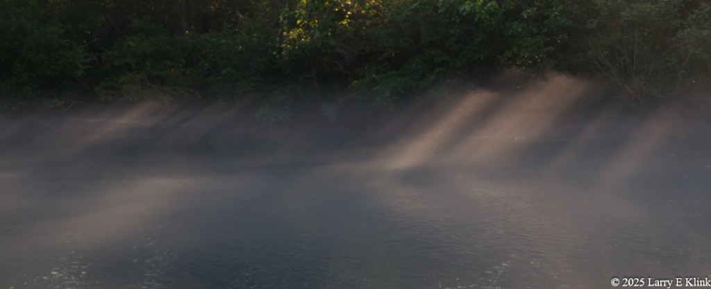 The picture is of a river with part of its far side bank. The river is covered by light fog. There are rays of sunlight filtering in through openings in trees along the far bank. The sunlight highlights portions of the fog creating shafts of light that illuminate and reflect from the river.