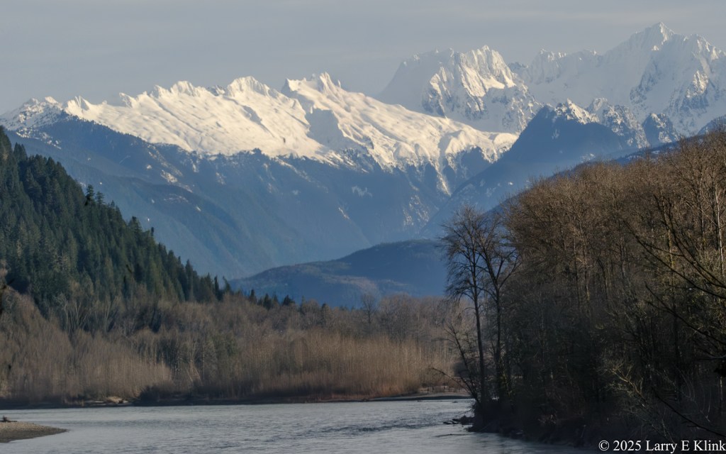 Snow-covered mountain peaks rise above layered blue foothills, a calm river flowing across the foreground and both leafless and evergreen forest lining the valley under a hazy sky.